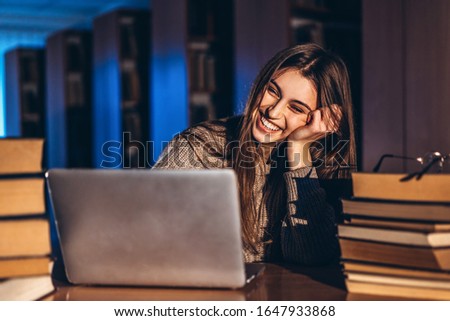 Young smiling student woman in the evening sits at a table in the library with a pile of books and works on a laptop. Preparing for the exam.