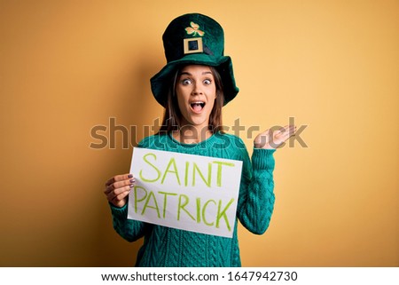 Woman wearing green hat celebrating st patricks day holding banner with saint patrick message very happy and excited, winner expression celebrating victory screaming with big smile and raised hands