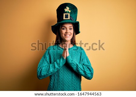 Young beautiful brunette woman wearing green hat with clover celebrating saint patricks day praying with hands together asking for forgiveness smiling confident.
