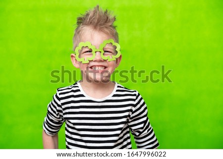 portrait little smiling caucasian boy in mask of leprechaun shamrock clover glasses for irish St. Patrick's Day on green studio background. Copyspace.