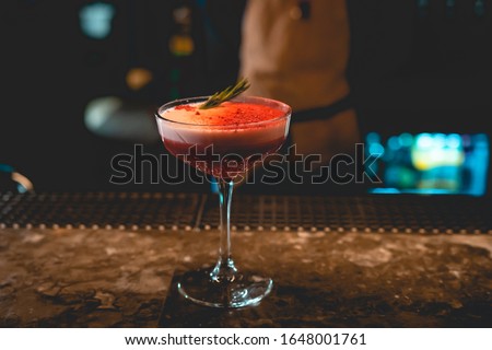 Clover Club cocktail with pink dried raspberries on the bar counter top made by male bartender with apron and tie looking sharp and fancy using hands pouring the alcohol in the glass  with rosemary up