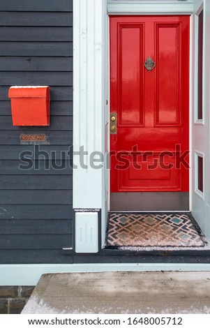 The entrance of a vintage home with blue wood siding, white trim and bright shiny red metal door with a brass knocker.  There's a mat on the doorstep of the building and a concrete step. 