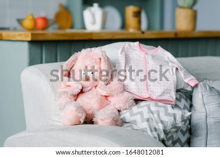 A baby blouse for a baby is lying on the sofa and a soft pink toy hare is sitting next to it.