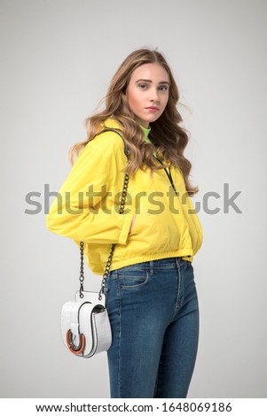 teenage blonde girl posing in a yellow raincoat with white crocodile leather bag and blue denim jeans. in studio on white background. 