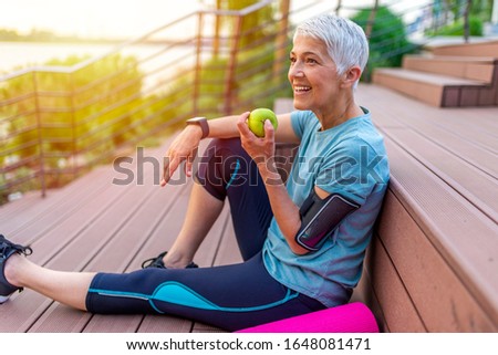 Portrait of a beautiful older woman with green healthy food after workout. Portrait of fit mature woman smiling while holding an apple and bottle of water. Sporty senior woman