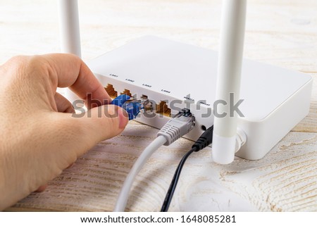 Woman hand plugging a blue network cable into socket of a white Wi-Fi wireless router on a white wooden table. Wlan router with connected internet cables on a table in a home or office. Rear view. Royalty-Free Stock Photo #1648085281
