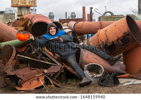 the worker lies on a pile of metal ducts and scrap metal. with a cheerful expression on his face holds a construction helmet in his hand. man has fun and jokes. Royalty-Free Stock Photo #1648119940