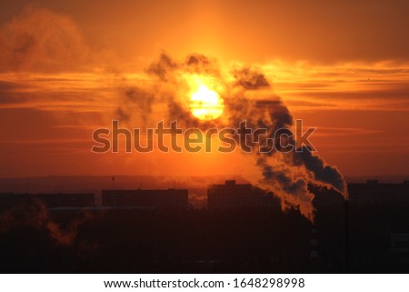 Red sun at sunset or dawn at dusk low above the horizon shrouded in smoke from the chimneys of the plant.