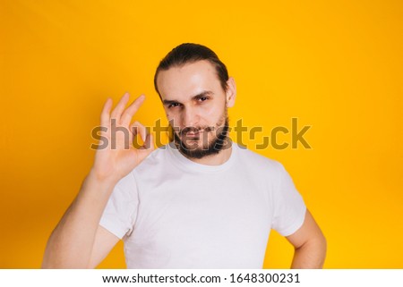 A guy with a beard in a white T-shirt on a yellow background shows a gesture well. Holds hand ok