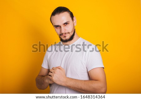 A guy with a beard in a white T-shirt on yellow folded his hands in the castle. Closed pose. Holding a fist