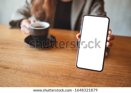 Mockup image of a businesswoman holding and showing black mobile phone with blank white screen while drinking coffee in cafe