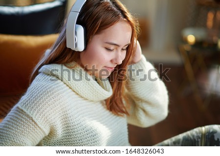 smiling modern young woman with red hair in white sweater listening to the music with headphones while sitting near couch at modern home in sunny winter day.