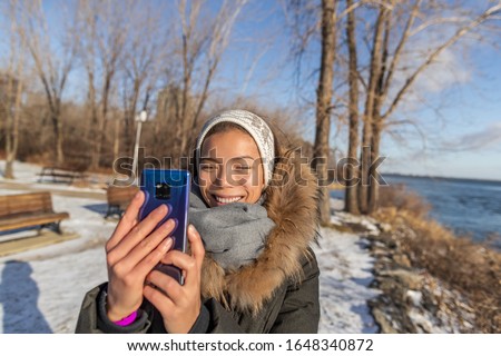 Winter travel Asian tourist woman taking selfie picture with mobile phone on nature forest trail walk holding smartphone with hands without gloves, cold weather frostbite care.