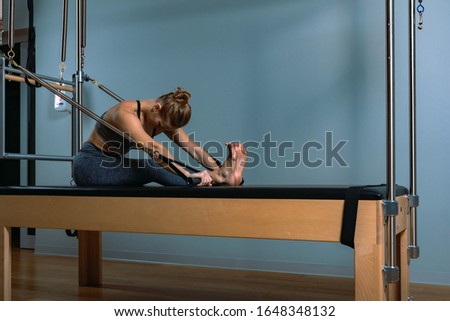 A young girl does Pilates exercises with a bed reformer, barrel machine tool. Beautiful slim fitness trainer on the background of a reformer doing various exercises gray, low key, light art. Fitness