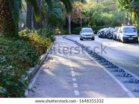 Bicycle path bordered by the lush vegetation of a park on the left and a one lane road with vehicles parked along and one car passing.