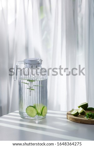 A clear glass jug with a drink with lime slices and balm is located on the white table near a cutting board with lime. The pitcher with a texture geometrical pattern is closed with a white cover. 