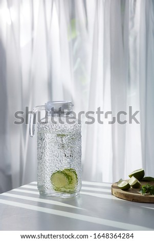 A clear glass jug with a drink with lime slices is located on the white table near a cutting board with lime and mint leaves. The pitcher with a texture pattern is closed with a white cover. 