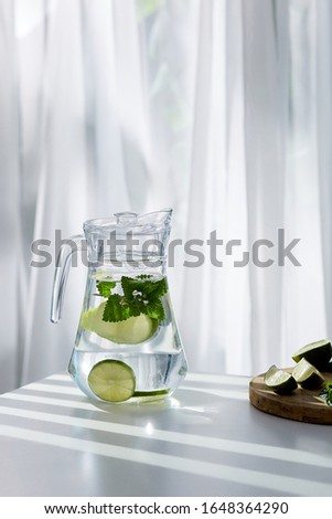 A clear glass jug with a drink with lime slices and balm is located on the white table near a cutting board with lime. The graceful pitcher is closed with a white cover. 