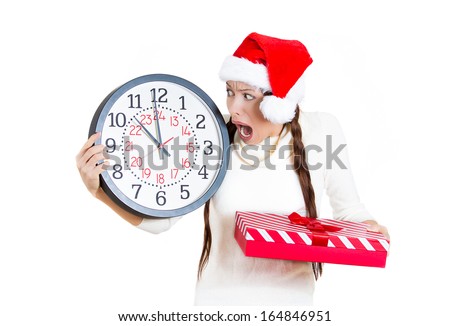 Closeup portrait worried stressed in a hurry young woman wearing red santa claus hat holding clock gift box isolated white background. Emotion, funny face expression, last minute christmas shopping