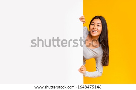 Pretty smiling asian girl looking from behind white advertising board over yellow background