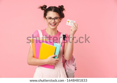 Photo of laughing nice girl in eyeglasses holding exercise books and credit card isolated over pink background