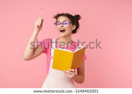 Photo of excited charming girl in eyeglasses writing in exercise book isolated over pink background