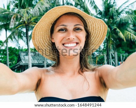 Close up shot of happy smiling female tourist in tropics enjoys free time outdoor near ocean on beach. Looks at camera during leisure on summer day. Poses for selfie