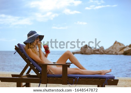 Beautiful young woman in bikini swimsuit sunbathes and rests on a wooden deck chair with mattress and red cocktail in glass with a straw