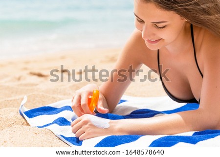 Cute and smiling woman is applying sunscreen on her hand with the finger sunbathing on the towel at the beach.