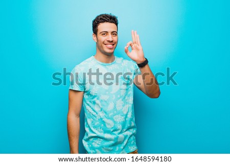 young arabian man feeling happy, relaxed and satisfied, showing approval with okay gesture, smiling against flat wall