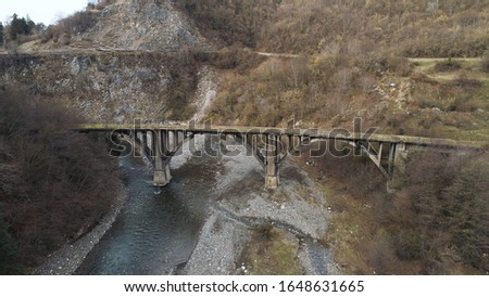 Flying under rusty bridge crossing the narrow river lwith stony bottom surrounded by mountains and forest. Shot. Aerial of rural landscape in late autumn.