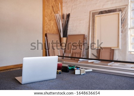Laptop, mat cutter and a variety of wooden picture frames on a table inside of a framing workshop