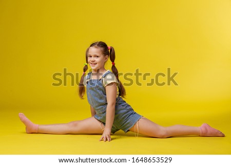 Girl schoolgirl in denim overalls is doing stretching. Sports modern dances leisure pastime. Yellow background place for text. Blond blue eyes preschooler. Child active positive cheerful smile workout
