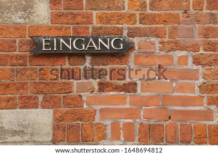 Black plate sign with metal letters and text translated from German "entrance" on a brick wall