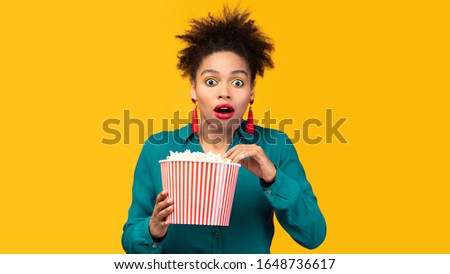 Wow. Surprised young black woman watching tv, holding popcorn over yellow wall, free space