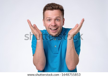 Excited casual man standing with raised hands and looking at camera with surprise. Studio shot. Positive facial human emotion.