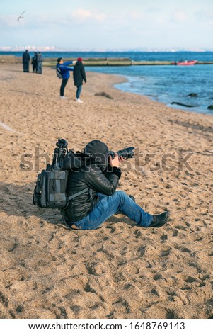Photographer traveler with a camera and a backpack on the beach in spring when it is still cold, takes pictures sea.