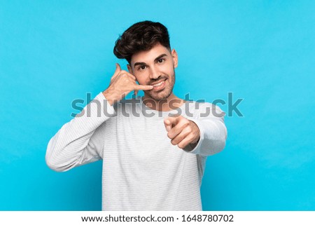 Young man over isolated blue background making phone gesture and pointing front