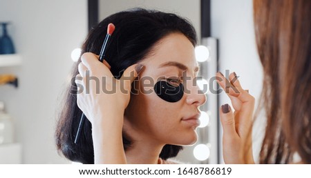 Portrait of brunette girl in makeup studio, makeup artist lays the foundation on skin with brush against background of bokeh light bulbs on mirror. Makeup and beauty. Lifestyle