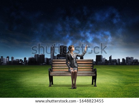 Young woman in business suit holding smartphone with raised hand. Beautiful girl with mobile phone on wooden bench. Mobile marketing and digital technology. Modern cityline panorama at night.