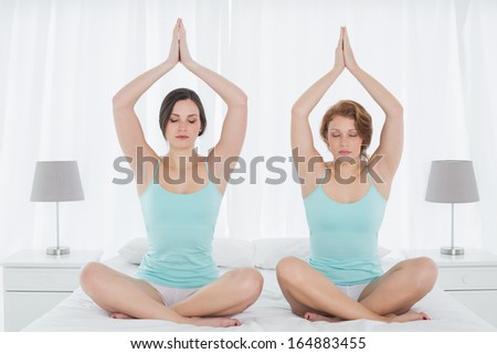 Full length of two young women sitting with joined hands over head on bed at home