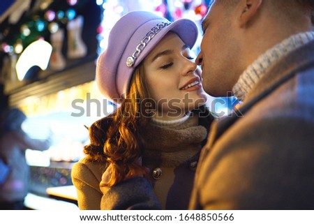 close-up portrait of romantic stylish couple in warm clothes close to each other and kissing, standing against ice rink with bright Cristmass tree. Spending time together at holidays, having a date
