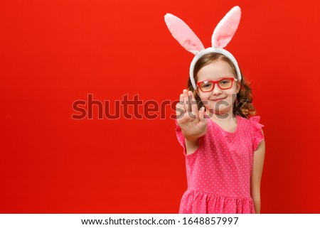 Little girl in easter bunny ears and glasses on a red background. The child shows a stop sign.
