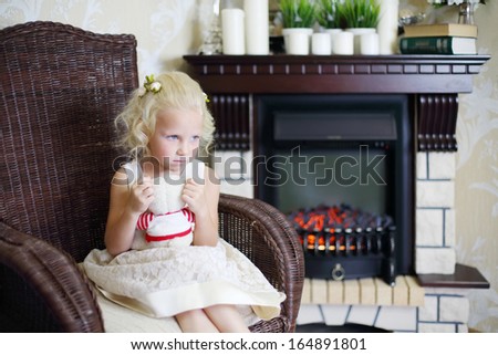 Little sad girl sits in wicker rocking chair with toy bear and looks away near fireplace in cosy room. 