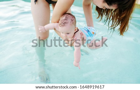 Insecure Caucasian 6 months old baby boy learning how to swim at swimming pool. Mother holding her son. First time at pool concept.