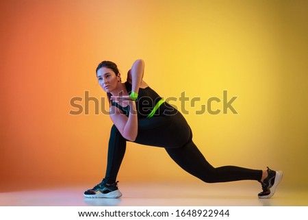 Young caucasian plus size female model's training on gradient orange background in neon light. Doing workout and stretching exercises. Concept of sport, healthy lifestyle, body positive, equality.