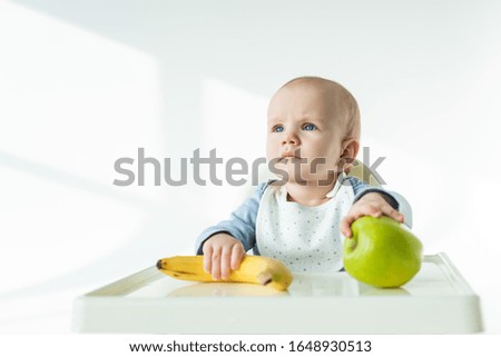 Adorable baby holding ripe banana and apple on table of feeding chair on white background