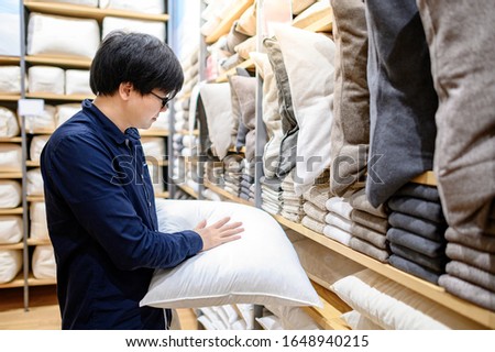 Young Asian man customer choosing throw pillow or sofa (couch) pillow in furniture store. Shopping house decoration for interior room design. Home improvement concept