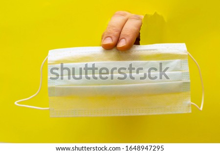 hand from a hole in a paper wall delivers a medical antivirus mask. yellow background
