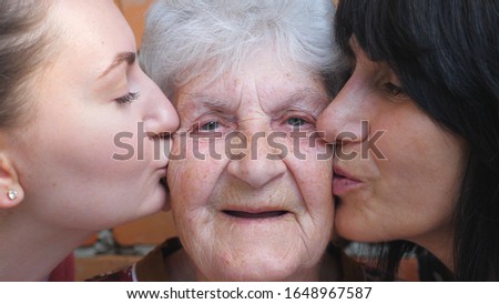 Young girl and adult woman kissing their beloved grandmother on cheeks. Portrait of happy granny smiling and looking into camera. Warm family relationships. Concept of love or care. Slow motion